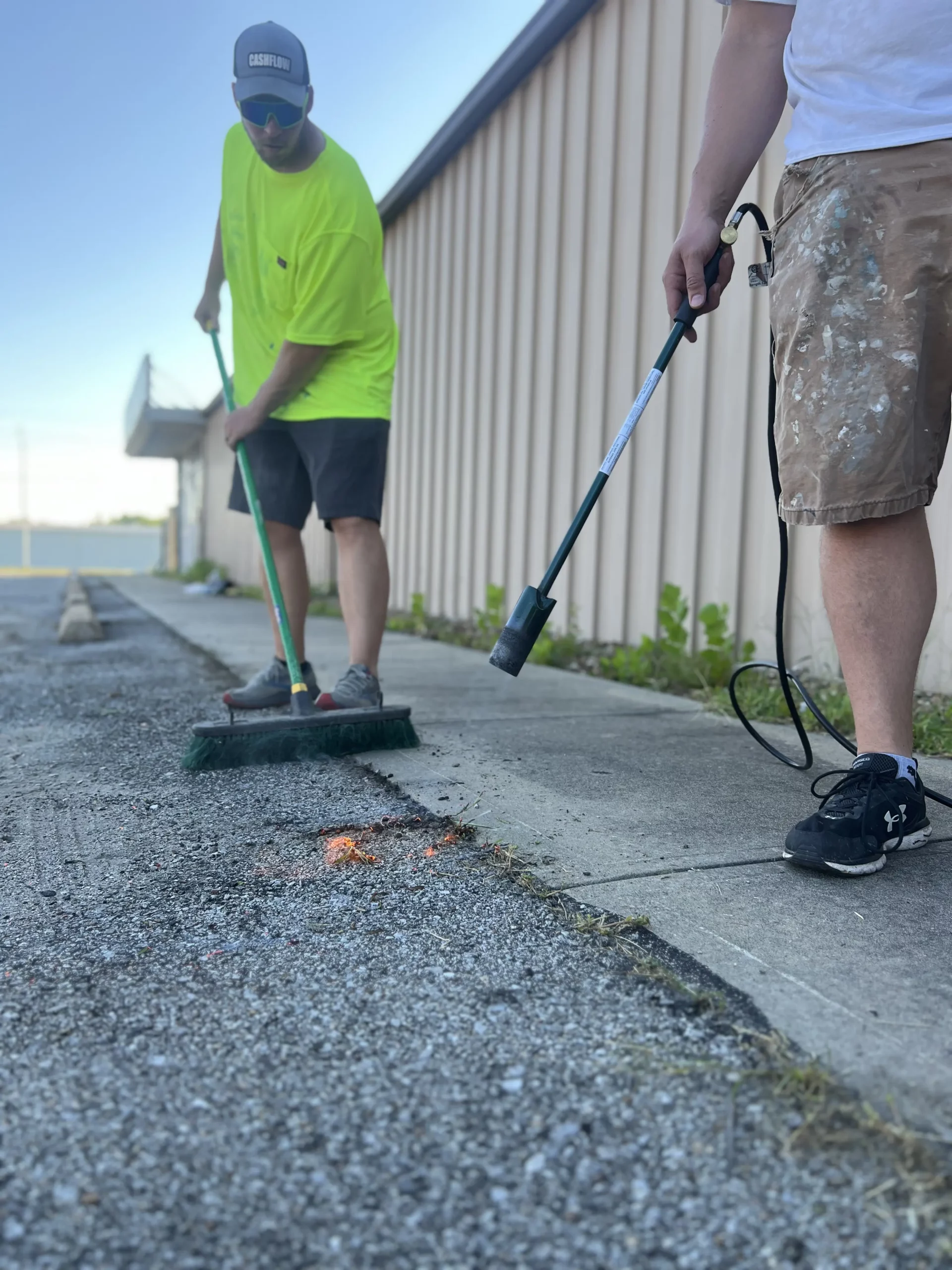 Workers burning weeds off a sidewalk and then scraping them off right after.