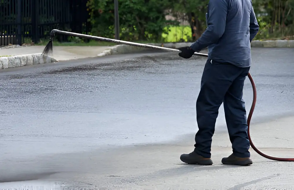 A worker wearing protective clothing spraying asphalt sealant on a parking lot surface.